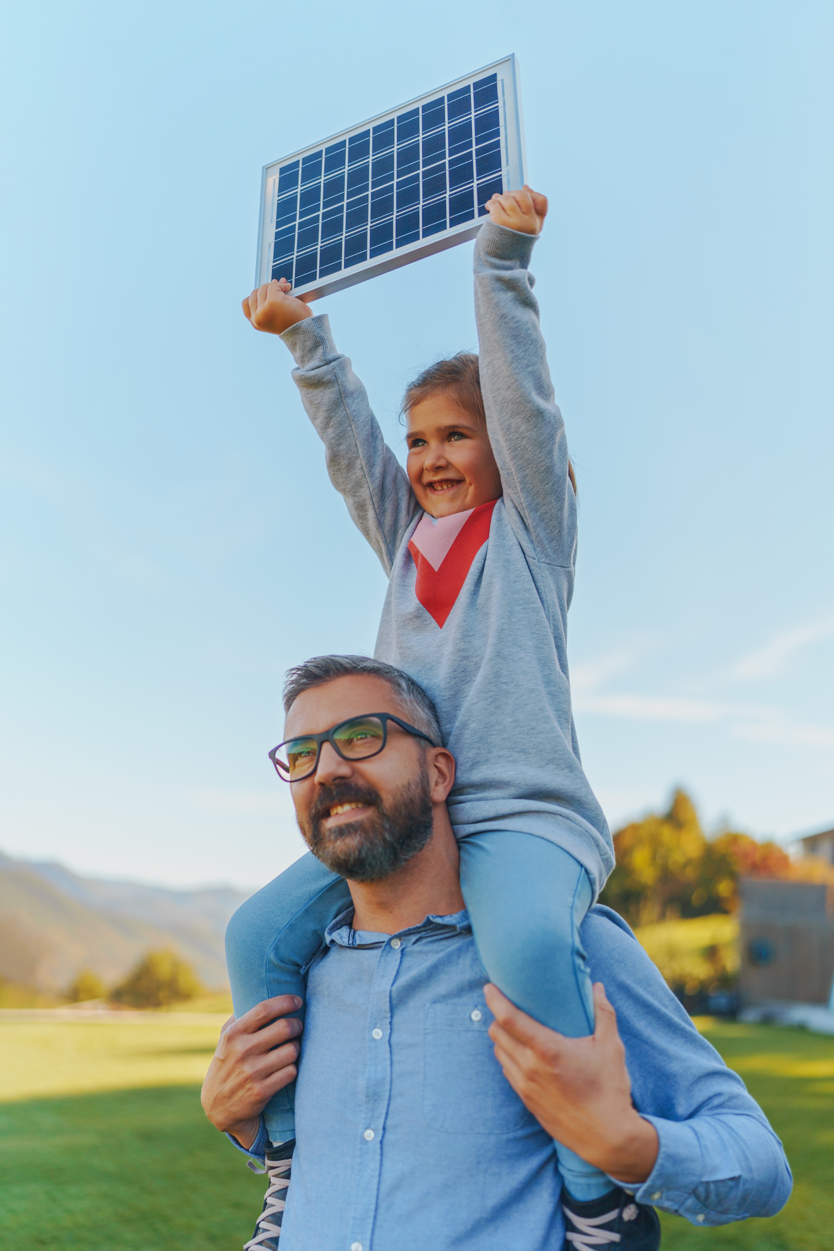 Father with his little daughter on piggyback, catching sun at solar panel,charging it at their backyard. Alternative energy, saving resources and sustainable lifestyle concept.